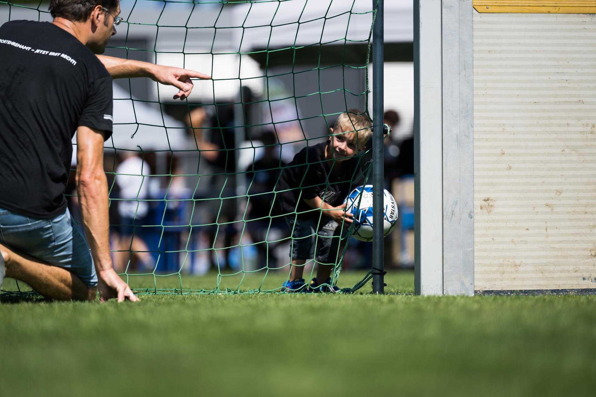 Ein Kind schaut durch ein Fußballtor, während ein Erwachsener auf es zeigt und ein Ball in der Hand hält.