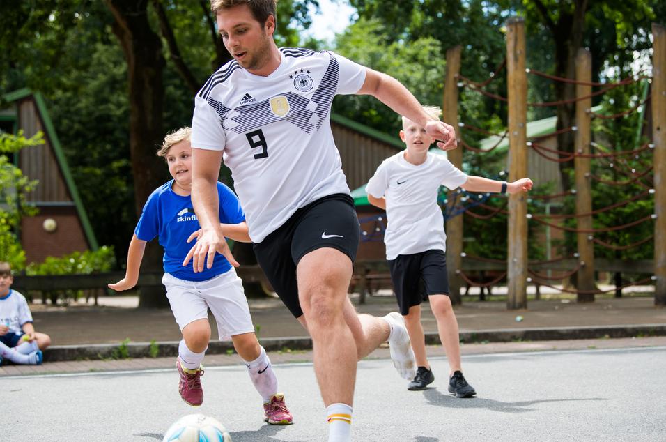Ein junger Mann in Fußballtrikot läuft mit einem Ball, während zwei Kinder ihm hinterherlaufen. Spielplatz im Hintergrund.